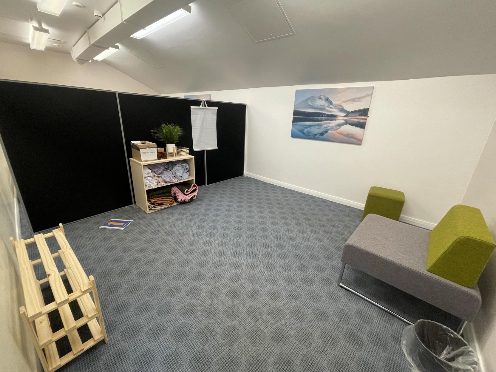 A simple room with grey carpet containing a comfortable chair, a wooden rack for shoes, and shelves containing prayer mats.  There is a picture on the wall of a mountain reflected in a lake.  The vaulted ceiling continues past a black room divider that separates the room into two halves.